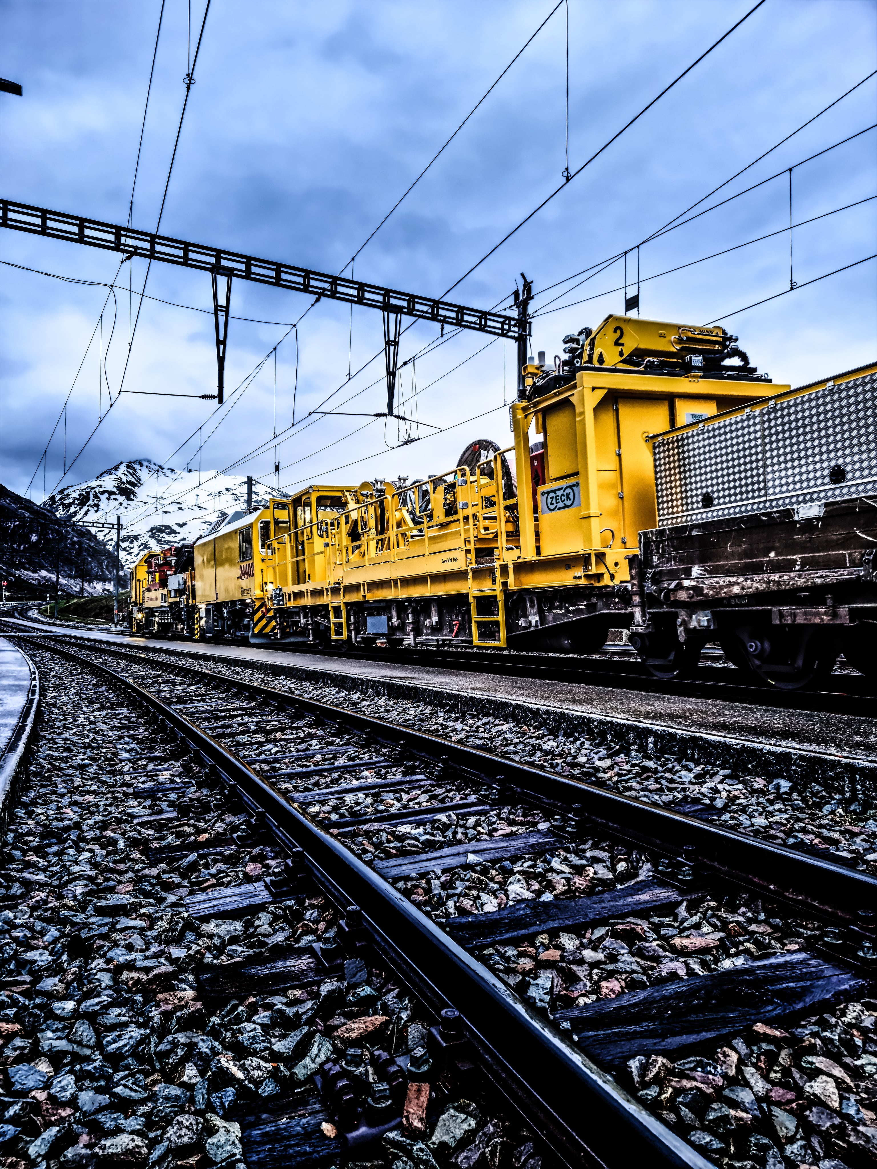 Gelber Arbeitszug mit einer ZECK-Einheit auf einem Waggon unter einer Oberleitung im Gebirge